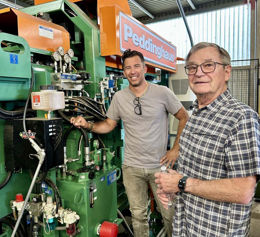 Two smiling men stand in front of an industrial machine